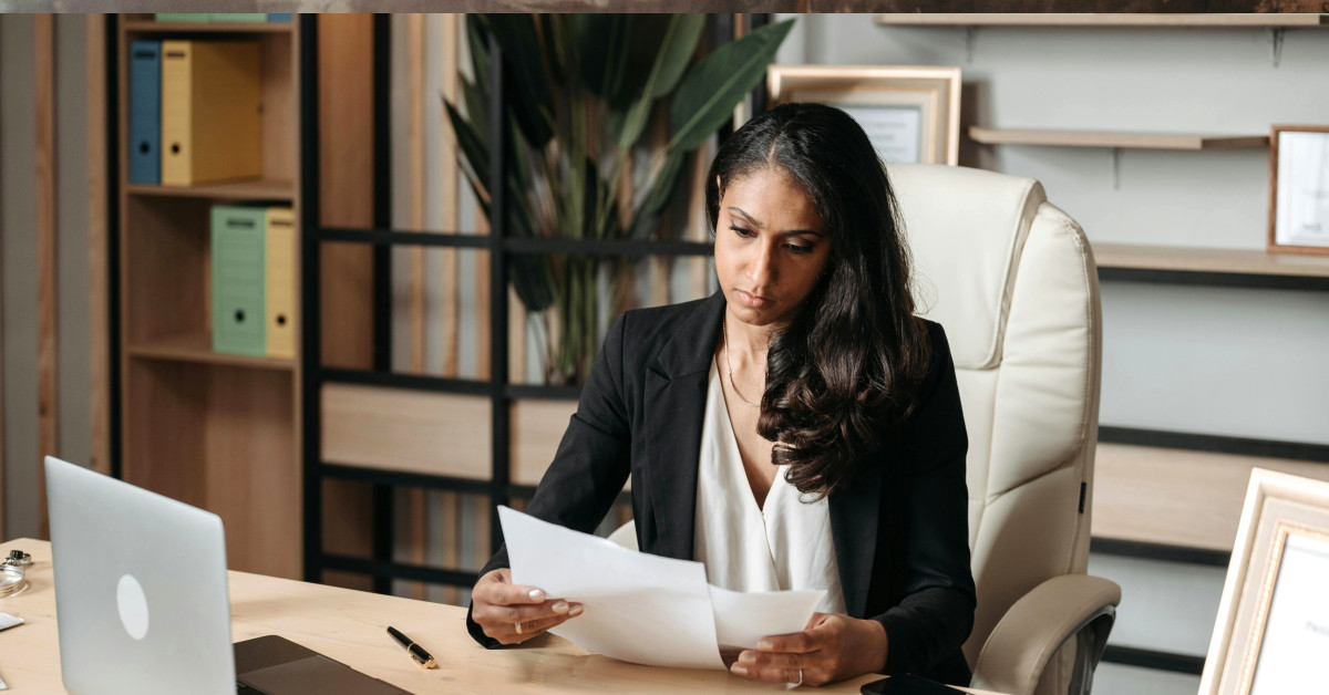 vrouw kijkt bedenkend naar papierwerk voor een laptop
